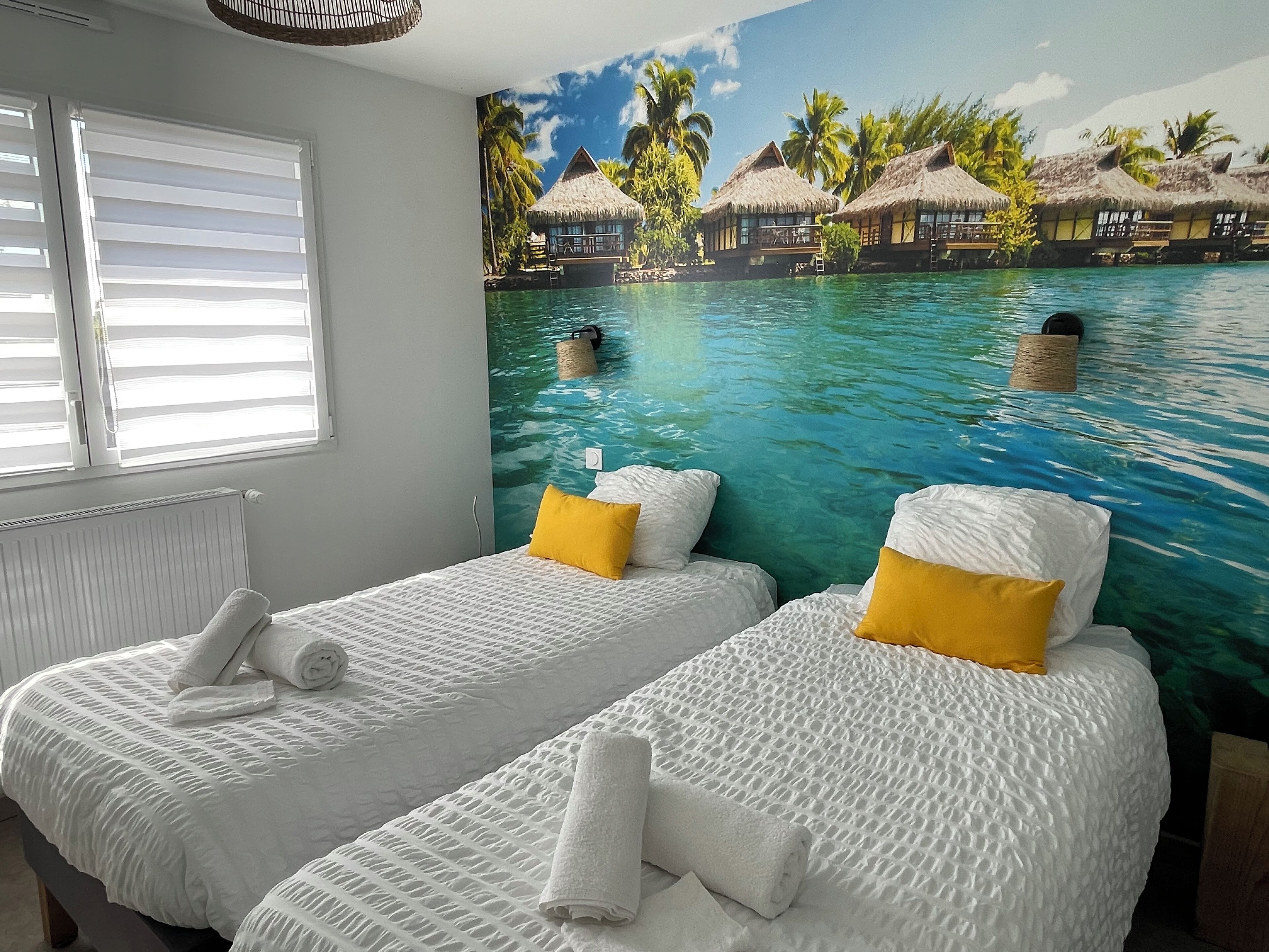 Une chambre avec deux lits, chacun doté de draps blancs et d’un oreiller jaune, et un mur décoré d’une fresque représentant un paysage de plage tropicale.