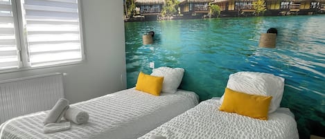 A room with two beds, each with white bedding and a yellow pillow, and a mural of a tropical beach scene.