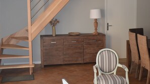 Una escalera que lleva a un nivel superior, un armario de madera con cajones, dos sillones tapizados, una lámpara y una puerta en un cuarto con suelo de baldosas.
