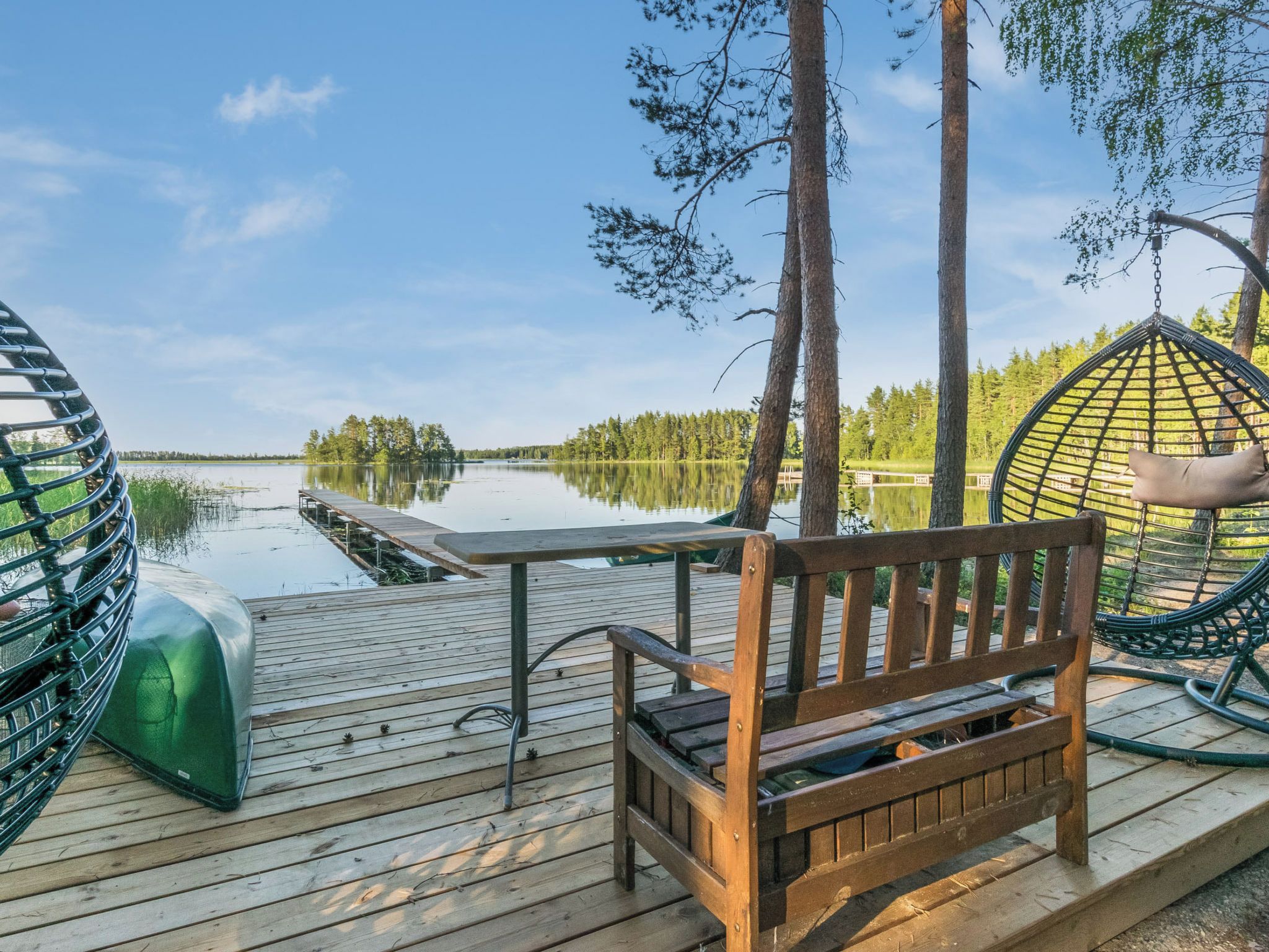 Eine Holzterrasse mit einer Bank und einem Hängesessel mit Blick auf einen ruhigen See, umgeben von Bäumen.