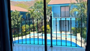 A room with a large window overlooking a swimming pool and a building with a red-tiled roof. - Charming Condo Orchidea (Playas Del Coco)