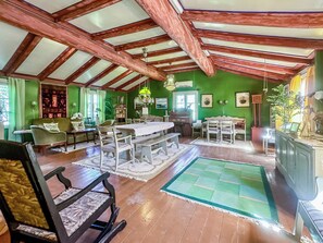 Een ruime kamer met een hoge plafond, houten balken, een eettafel, stoelen, een bank en een schommelstoel.
