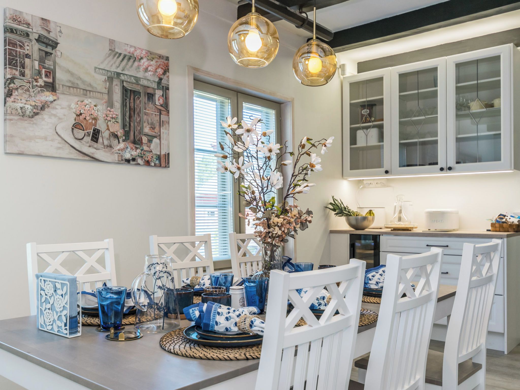 Una cucina moderna con mobili bianchi, un tavolo da pranzo apparecchiato con stoviglie blu e bianche, e un grande quadro floreale appeso al muro.