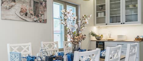 A modern kitchen with white cabinetry, a dining table set with blue and white tableware, and a large floral painting on the wall.