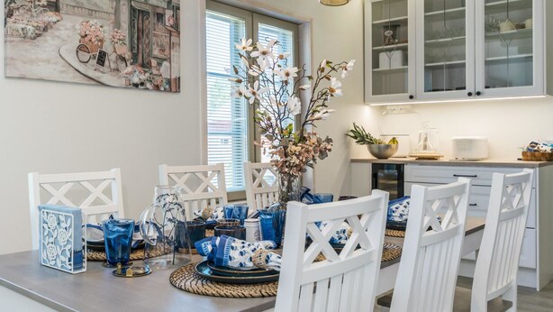 A dining area with a long table, white chairs, and a vase of flowers. There are hanging lights above the table and a painting on the wall.