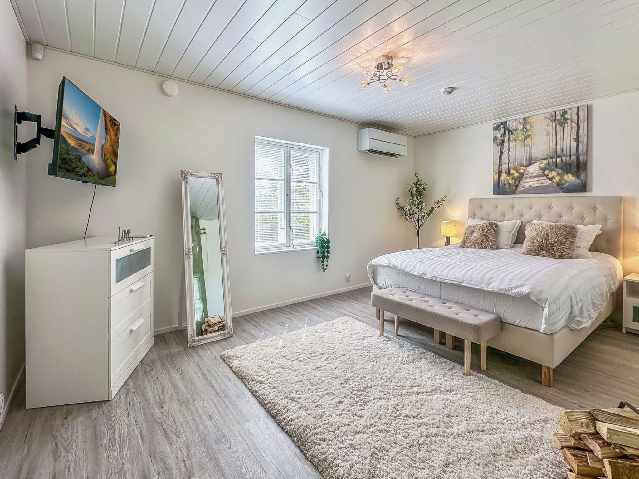 Une chambre à coucher avec un lit, des tables de chevet, un miroir, une commode, une fenêtre et un tableau accroché au mur.