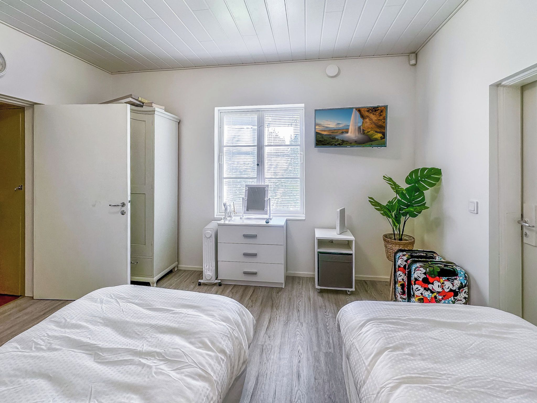 A bedroom with two beds, a window with blinds, a desk with a chair, a radiator, and a plant.