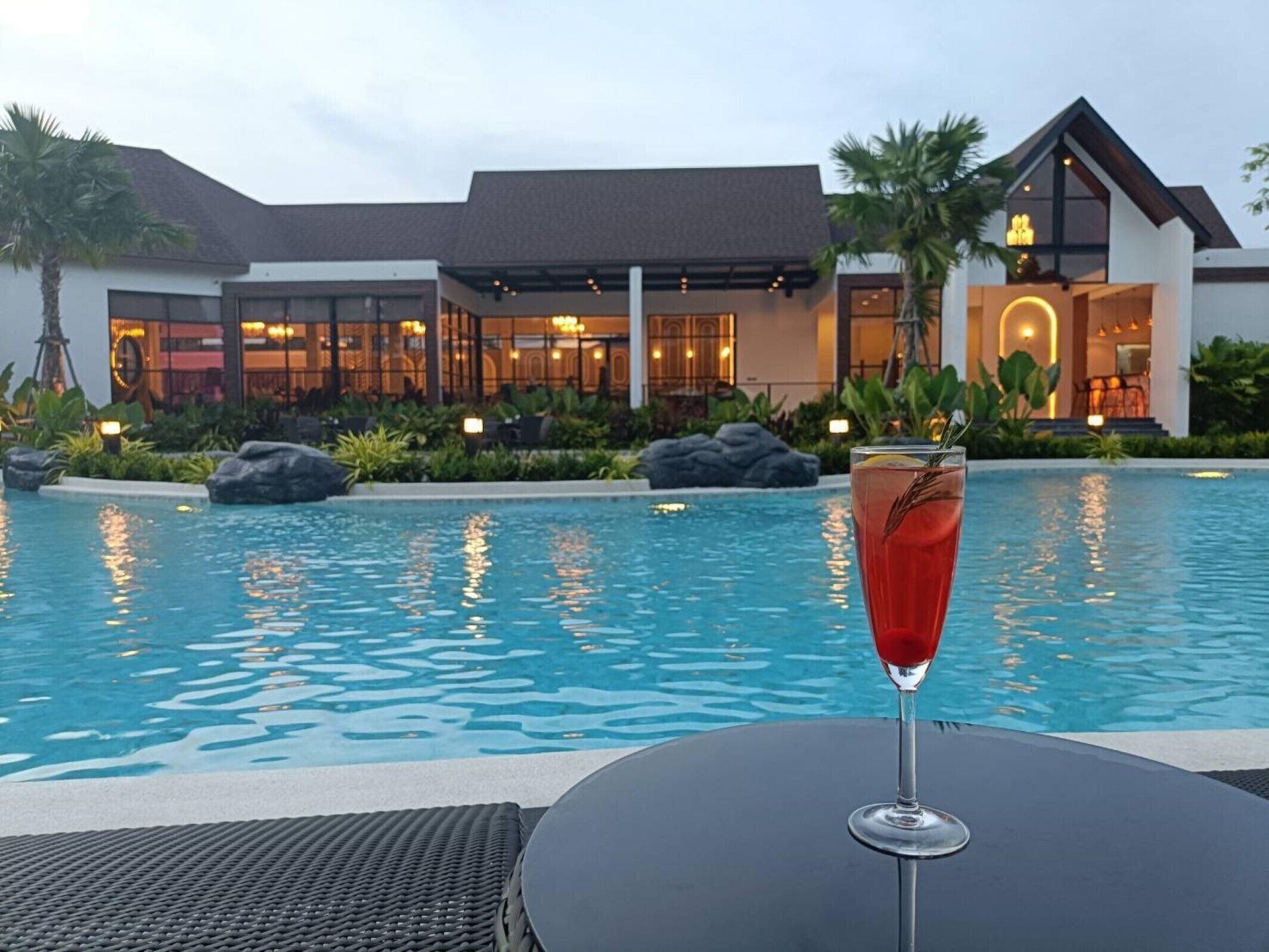 Uitzicht op het zwembad met een glas rode drank, een ligstoel, en een gebouw met modern design, met een restaurant in de achtergrond.