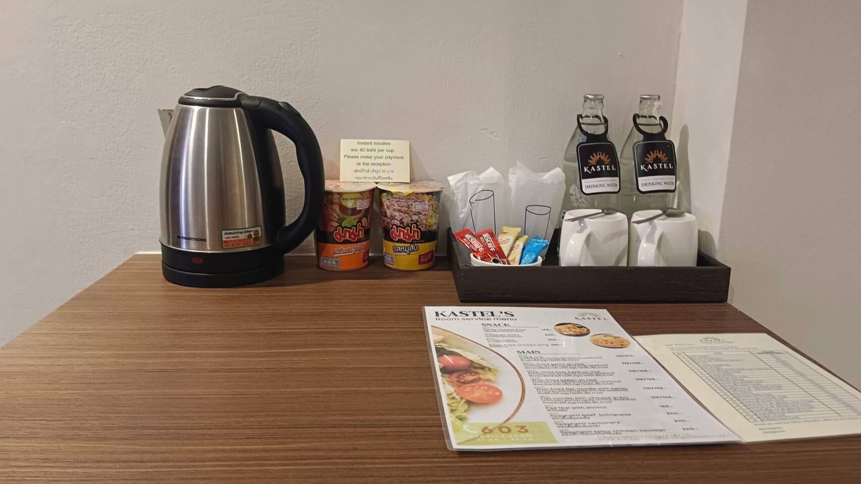 A wooden table with a stainless steel kettle, instant noodles, two bottles of Kasteel drinking water, a napkin holder, and a room service menu.