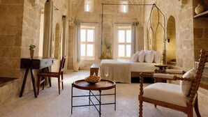 Une pièce spacieuse avec des arches en pierre, un lit, une chaise en bois, une table sur laquelle se trouve un saladier rempli de fruits, et un luminaire suspendu.