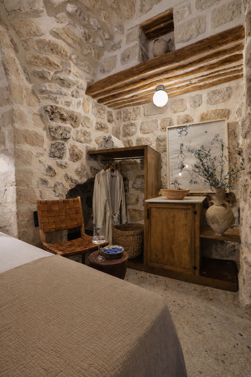 Une pièce rustique aux murs de pierre, avec une chaise en bois, une petite table sur laquelle se trouve un verre, et une étagère ornée d’un vase et d’un bol.