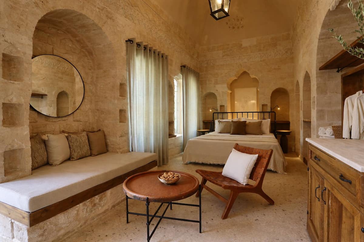 A room with a stone wall, a bed with a headboard, a wooden bench, a round table, and a mirror.