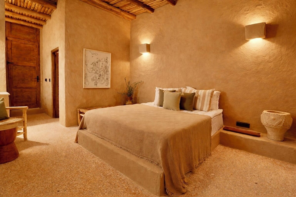 A bedroom with a large bed, a wooden door, a framed artwork, and a stone vase.