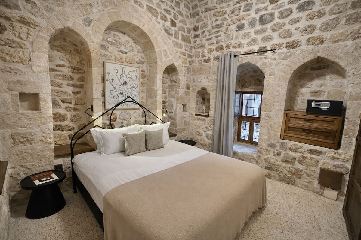 A stone-walled bedroom with a bed, a bedside table, a nightstand, a framed artwork, and a window with curtains.