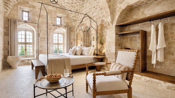 Une chambre aux murs de pierre, avec un lit, une chaise en bois, une petite table sur laquelle se trouvent un bol et un verre, et un luminaire suspendu au plafond.