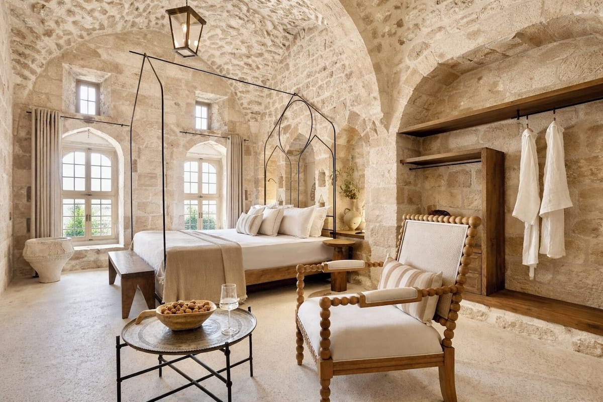 A stone-walled bedroom with a bed, a wooden chair, a small table with a bowl and glass, and a hanging light fixture.