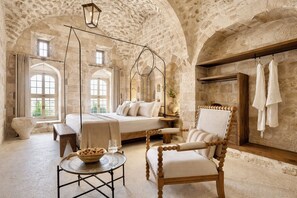 A stone-walled bedroom with a bed, a wooden chair, a small table with a bowl and glass, and a hanging light fixture.