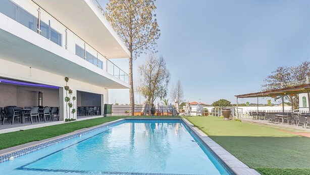 A rectangular swimming pool with a tiled edge, surrounded by a well-maintained lawn and a building with outdoor seating. - Botle Ba Afrika Lodge (Vereeniging)