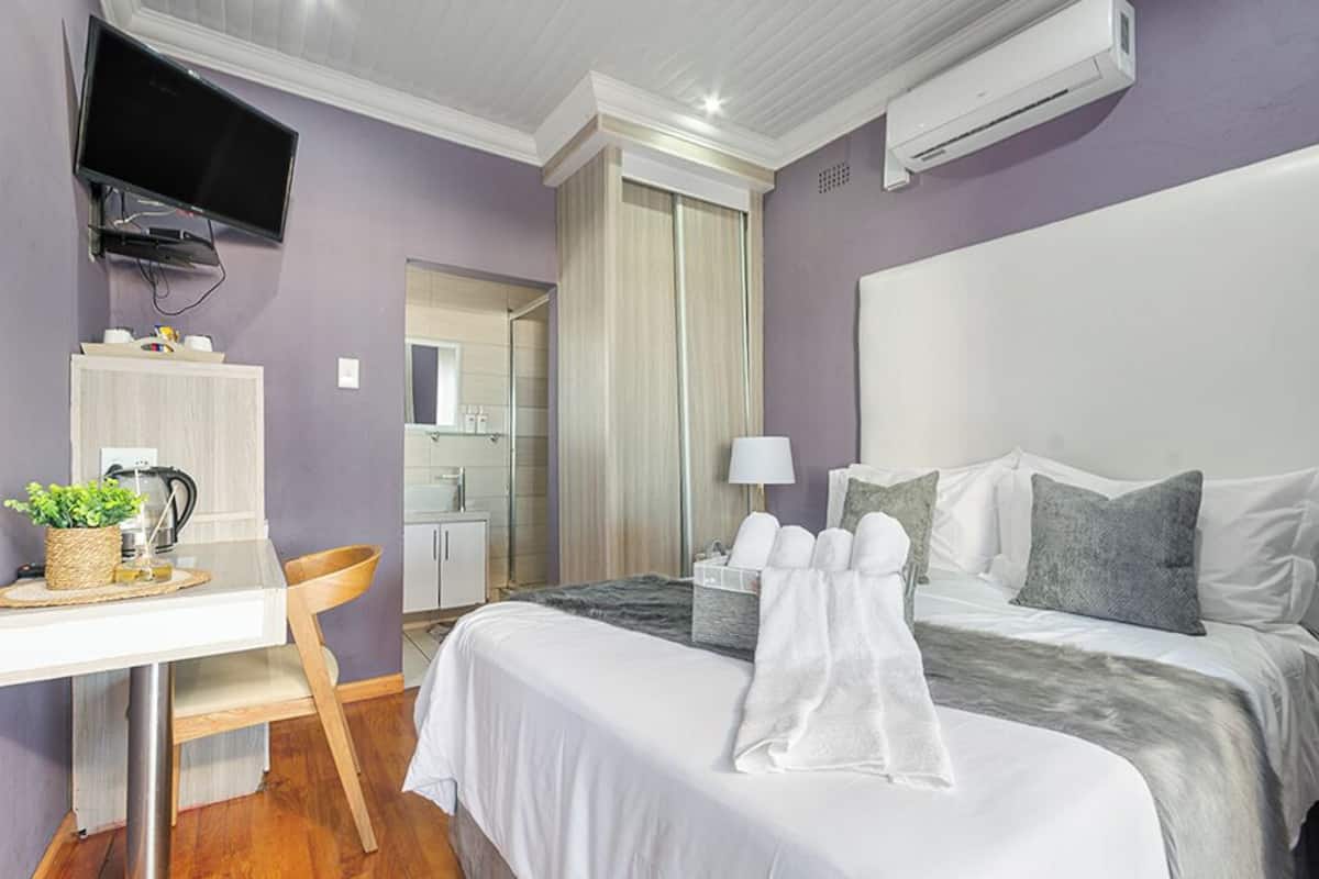 A hotel room with a bed, a desk with a chair, a TV mounted on the wall, and an air conditioning unit.