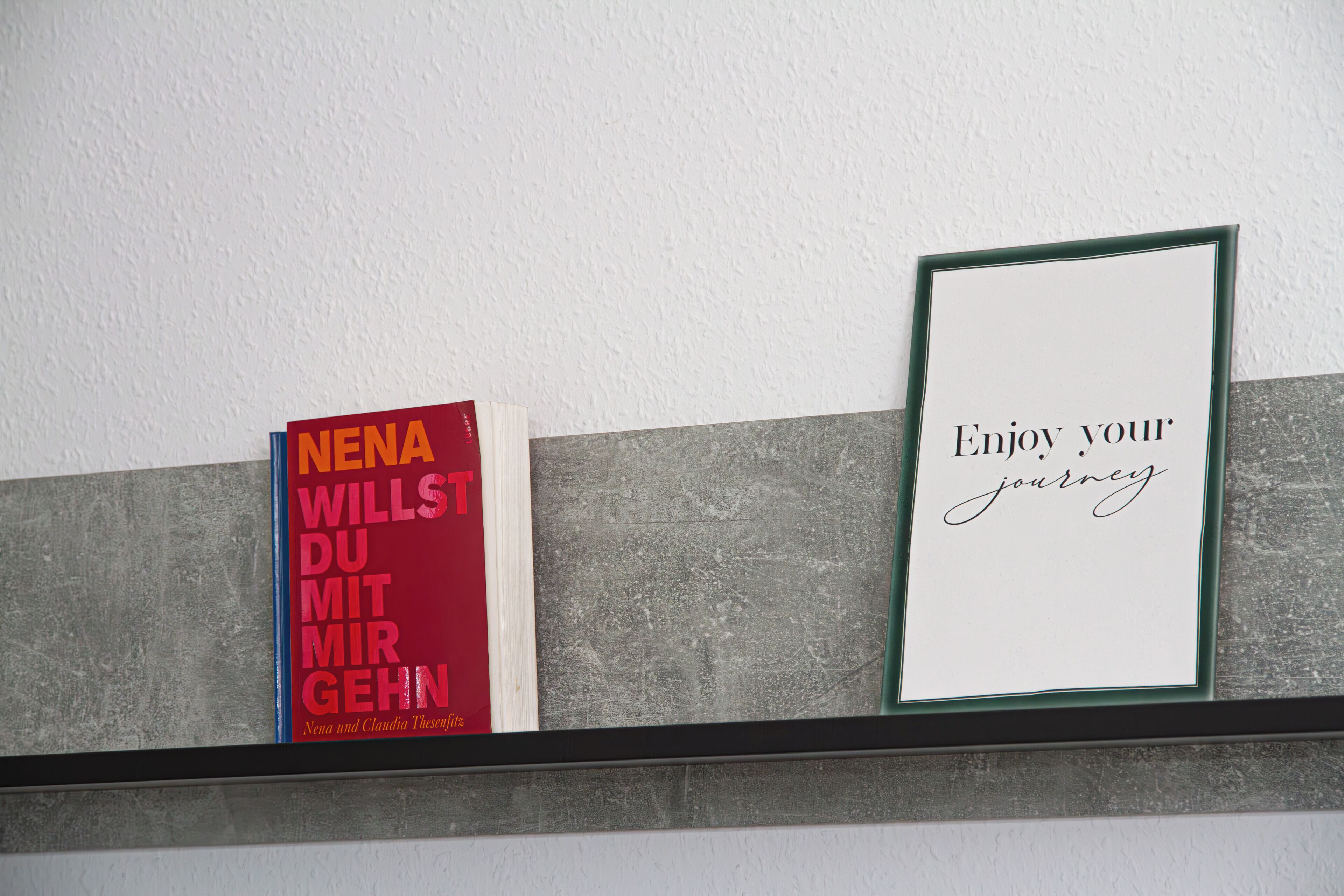 Ein Regal mit zwei gerahmten Gegenständen: ein Buch mit dem Titel 'NENA WILLST DU MIT MIR GEHNT' und ein gerahmter Spruch 'Genieße deine Reise'.