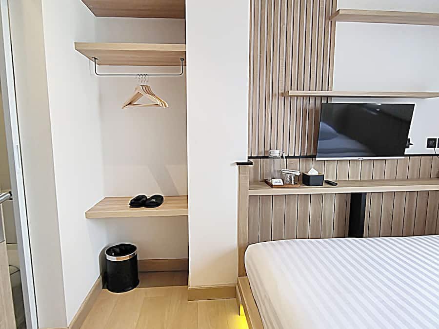 A modern hotel room with a bed, a TV mounted on the wall, a small shelf with a hanger, and a trash bin.