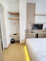 A modern hotel room with a bed, a TV mounted on the wall, a small shelf with a hanger, and a trash bin.