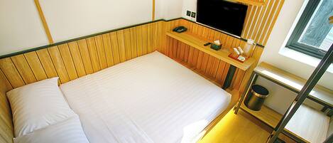 A single bed with white linens, a wooden headboard, a small bedside table with a television, and a window with blinds.