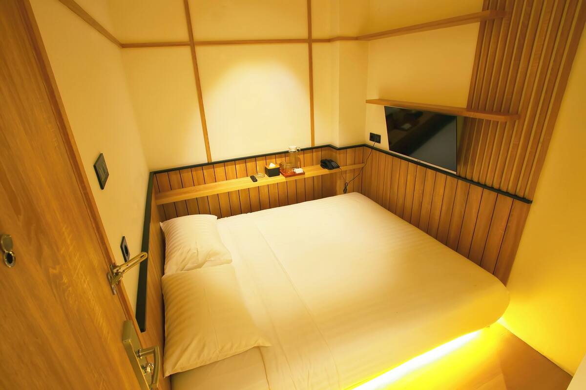A neatly made bed with a wooden headboard, a bedside table with a lamp, and a wall-mounted TV.