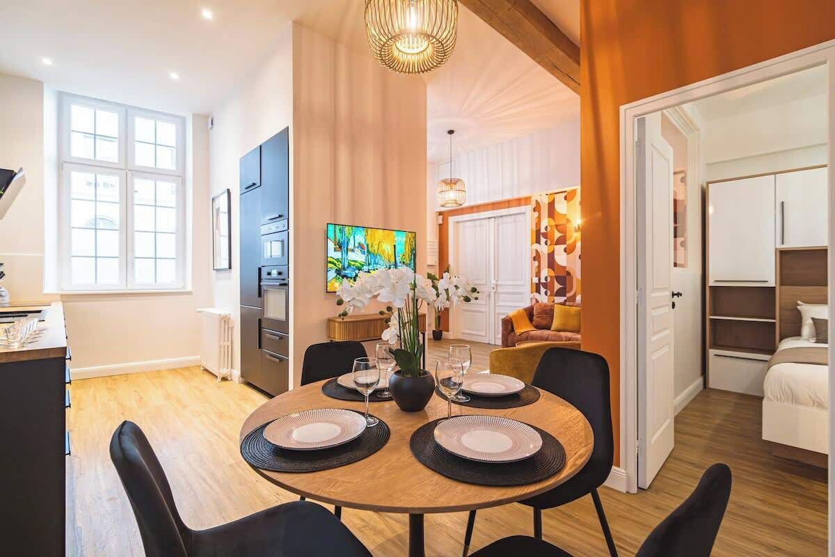 Un comedor moderno con una mesa redonda dispuesta para cuatro, una cocina al fondo y una sala de estar con sofá y un cuadro.