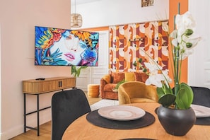 A dining area with a round table, chairs, a television mounted on the wall, and a patterned wall in the background. - MM Suites Loft Cathédrale (Reims)