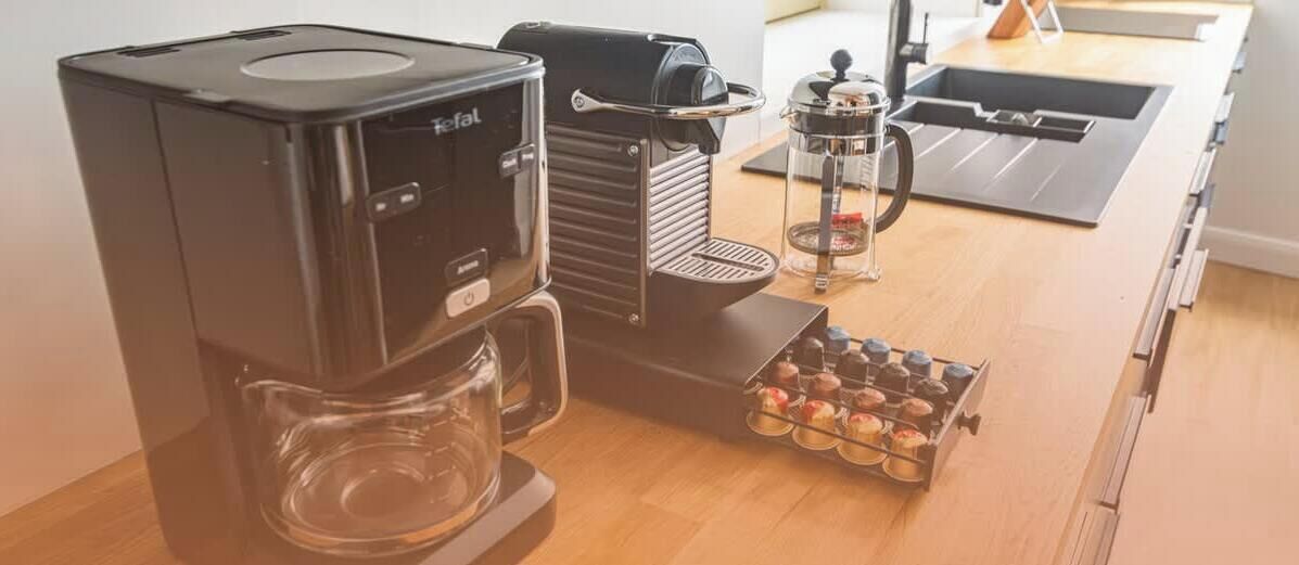 Kahvinkeitin, jossa on lasinen kannu ja kori, jossa on kahvikapseleita, ja ne ovat sijoitettu puiseen työtasoon.