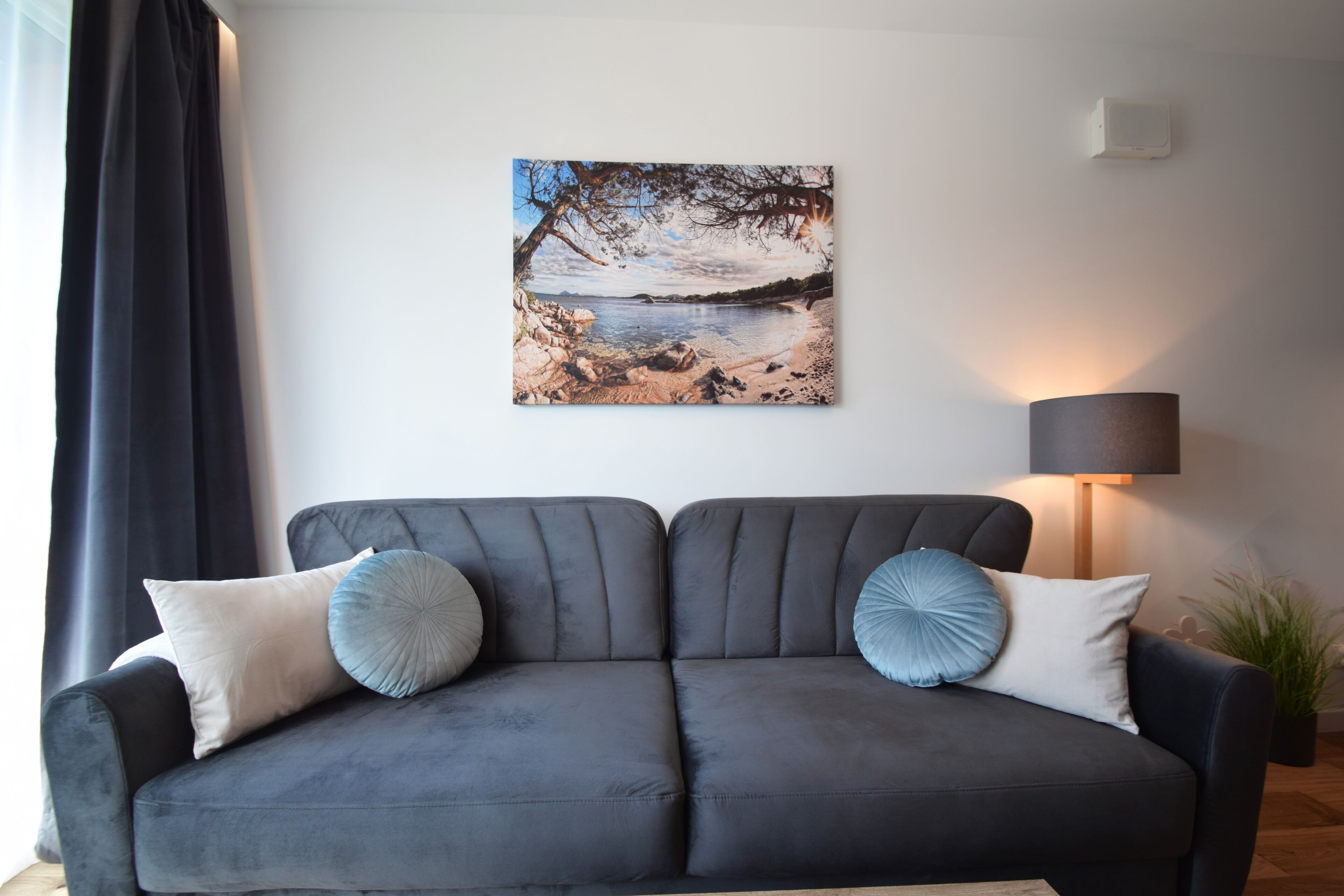 Ein modernes Wohnzimmer mit einem grauen Sofa, zwei Kissen, einem Wandbild mit Seeblick und einer Stehlampe.