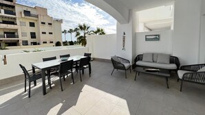 A terrace with a dining table and chairs, a sofa, and a small table. There is a view of buildings and palm trees in the background. - PUSHE Playa Granada Beach & Golf 12 (Motril)