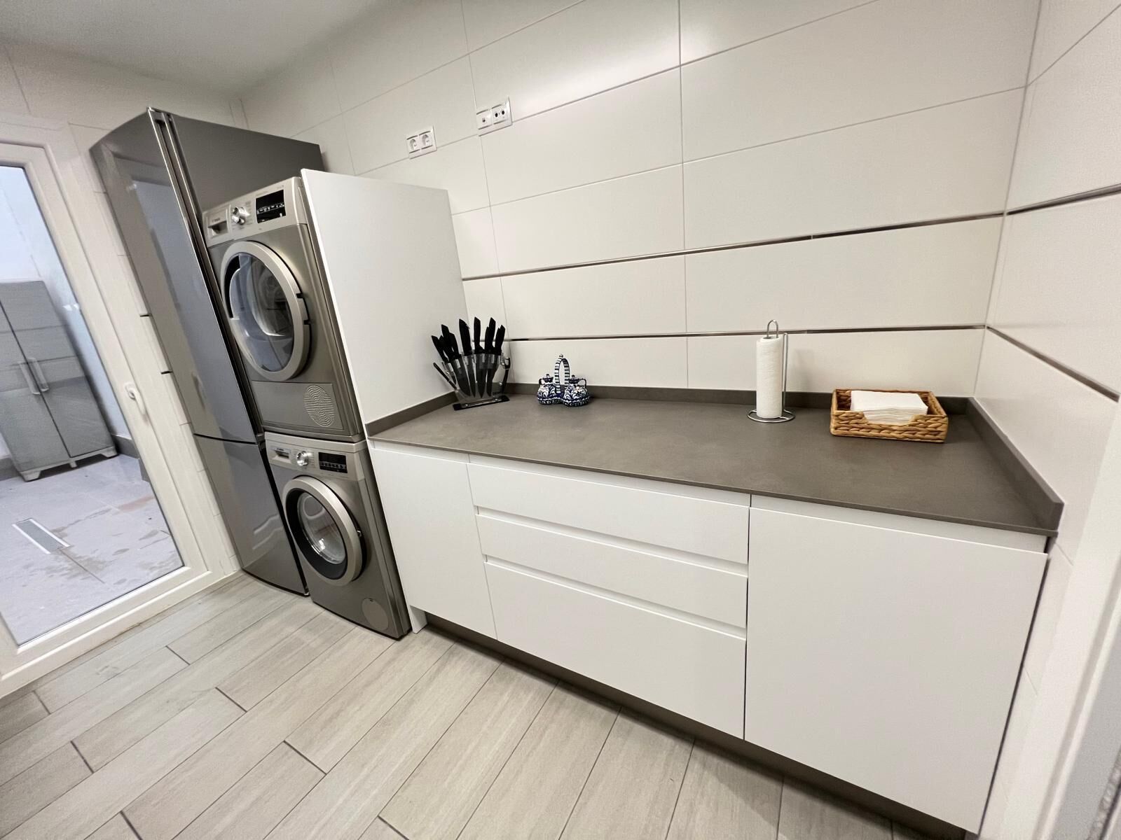 A modern kitchen with a built-in washer and dryer, white cabinetry, a grey countertop, and a small wicker basket.