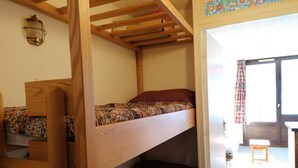 Ein Schlafraum mit Etagenbetten, einer Holzlade, einem kleinen Tisch und einem Fenster mit Vorhängen.