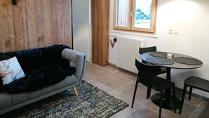Un salon chaleureux avec un canapé bleu foncé, une petite table ronde entourée de chaises, un radiateur et une fenêtre avec des cadres en bois.