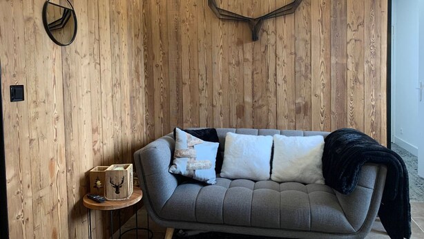 Une pièce chaleureuse avec un canapé gris, des murs lambrissés, une tête de cerf accrochée au mur et une petite table d’appoint en bois sur laquelle repose une télécommande.