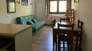 A cozy room with a green sofa, wooden dining table, and chairs. There are two paintings on the wall and a window with curtains. - Cozy studio for 2 near slopes in Pra-Loup (Pra-Loup)