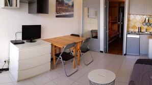 Un espacio habitable compacto con una mesa de comedor de madera, una mesita redonda pequeña, un televisor y una kitchenette.