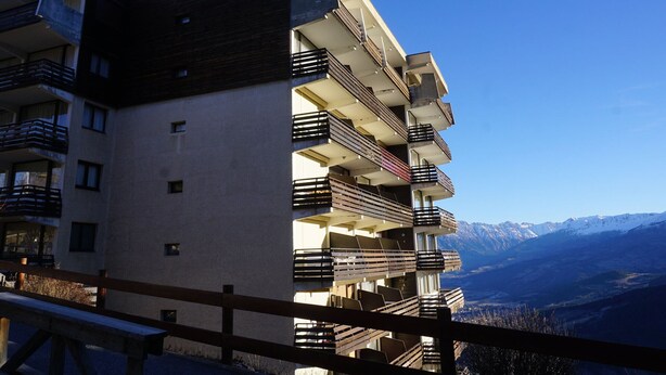 Un edificio de varios pisos con balcones, una valla de madera en primer plano y un paisaje montañoso al fondo.