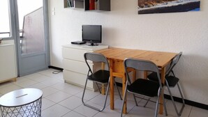 Un pequeño comedor con una mesa de madera, cuatro sillas, un televisor sobre un mueble blanco y un cuadro de un paisaje nevado en la pared.