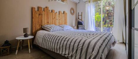 A bedroom with a wooden headboard, a bed with striped bedding, a small round table with a lamp, a ladder, and a window with curtains.