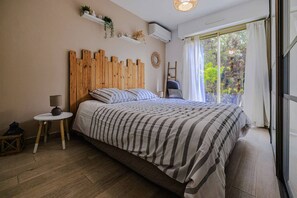 Un dormitorio con cabecera de madera, cama con ropa de cama a rayas, una pequeña mesa redonda con una lámpara, una escalera y una ventana con cortinas.