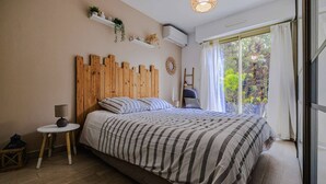A bedroom with a wooden headboard, a bed with striped bedding, a small round table with a lamp, a ladder, and a window with curtains. - Marina Rey – 400m from beach, Côte d’Azur vibe (Villeneuve-Loubet)