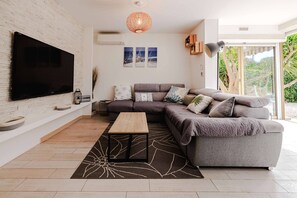 A modern living room with a large flat-screen TV, a sectional sofa, a coffee table, and a large window with a view of greenery. - Marina Rey – 400m from beach, Côte d’Azur vibe (Villeneuve-Loubet)