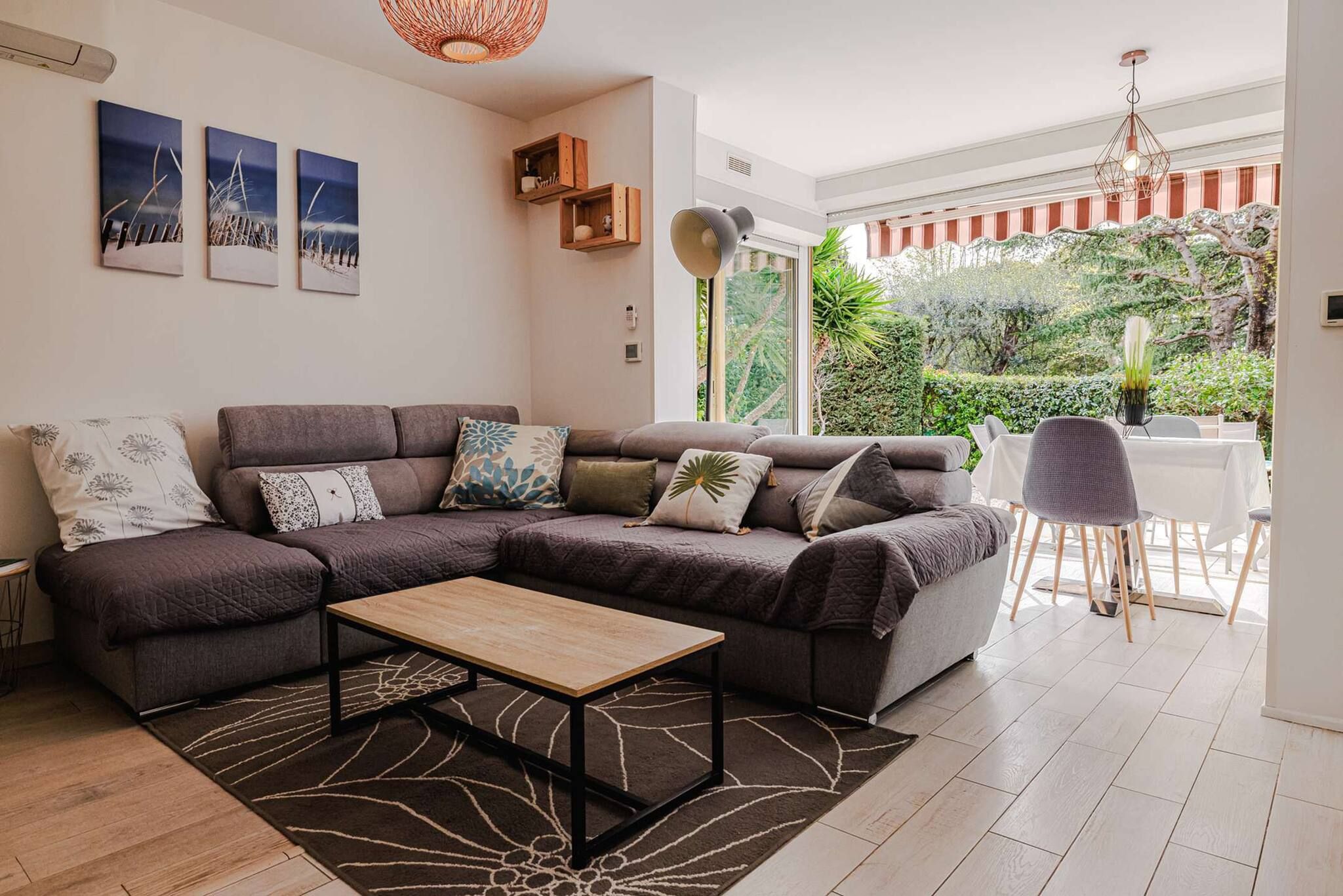 A modern living room with a sectional sofa, a coffee table, and a dining area with a table and chairs. There are three wall-mounted artworks and a hanging light fixture.