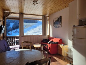 A cozy room with a wooden ceiling, a large window with a snowy mountain view, a red sofa, a wooden table, and a bed with a purple blanket.