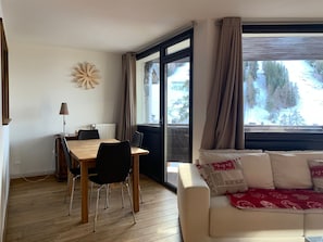 A modern living room with a wooden dining table, black chairs, a beige sofa with patterned cushions, and a large window with curtains. - Renovated 4 pers apt, balcony, close to slopes, parking (Pra-Loup)