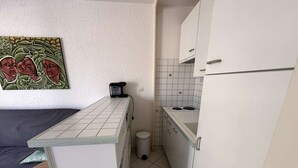 A compact kitchen with a small countertop, a stove, and a trash bin. A painting is visible on the wall above the countertop. - Charming Studio for 4 with Sea View, Pool, and Parking Near the Beach (Le Grau-du-Roi)