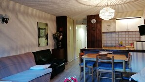 A cozy living room with a sofa, a dining table with chairs, a kitchen area, and a decorative vase with flowers. - South-facing studio with balcony, parking, 4 people (Pra-Loup)